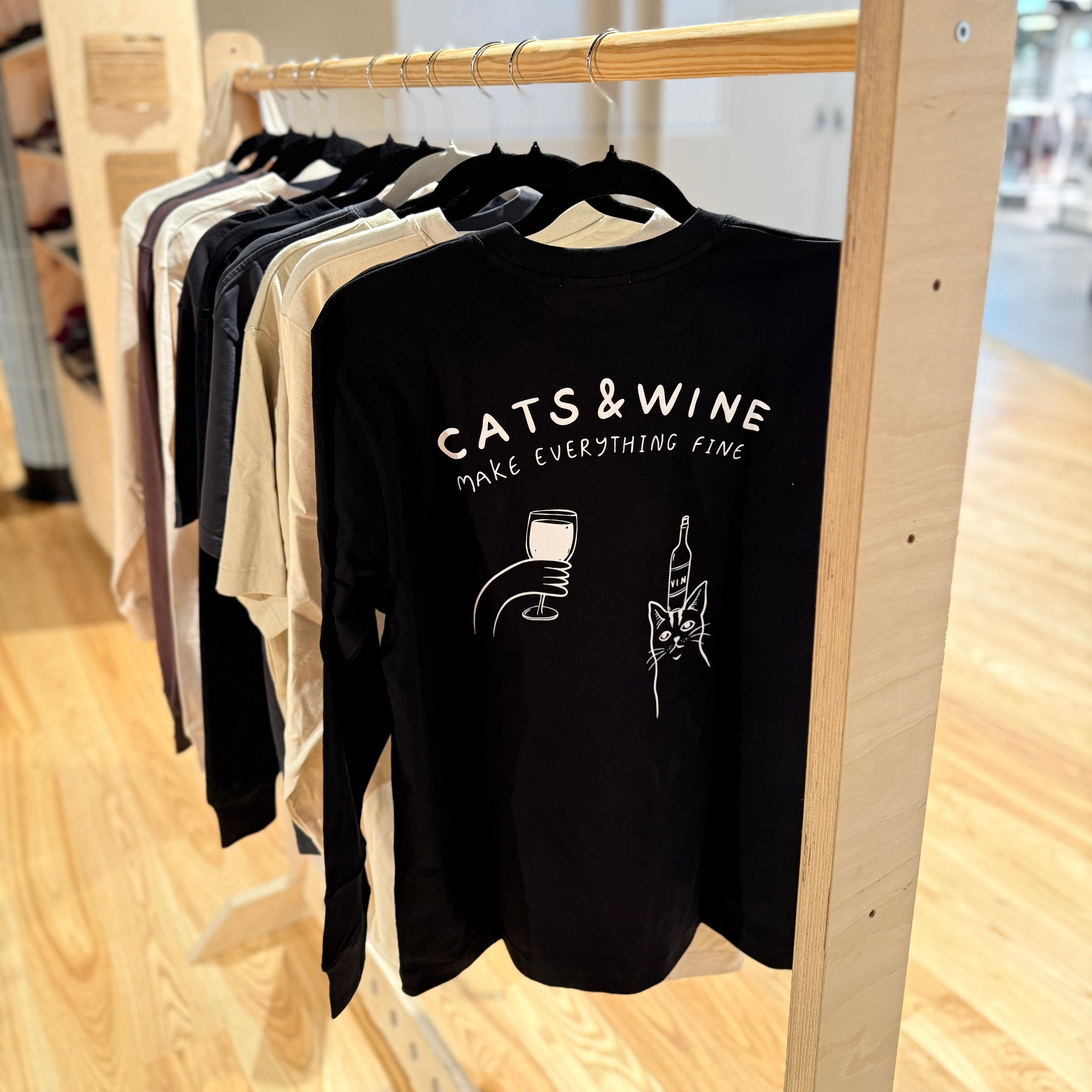 Black sweatshirt with 'Cats & Wine Make Everything Fine' text and graphics on a wooden rack.